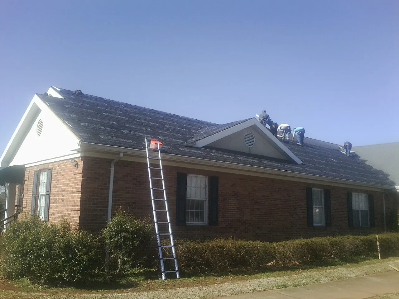 Crew tearing shingles off a brick ranch roof with a ladder set — Metal Roof Repair work by our crew in Clifton Springs