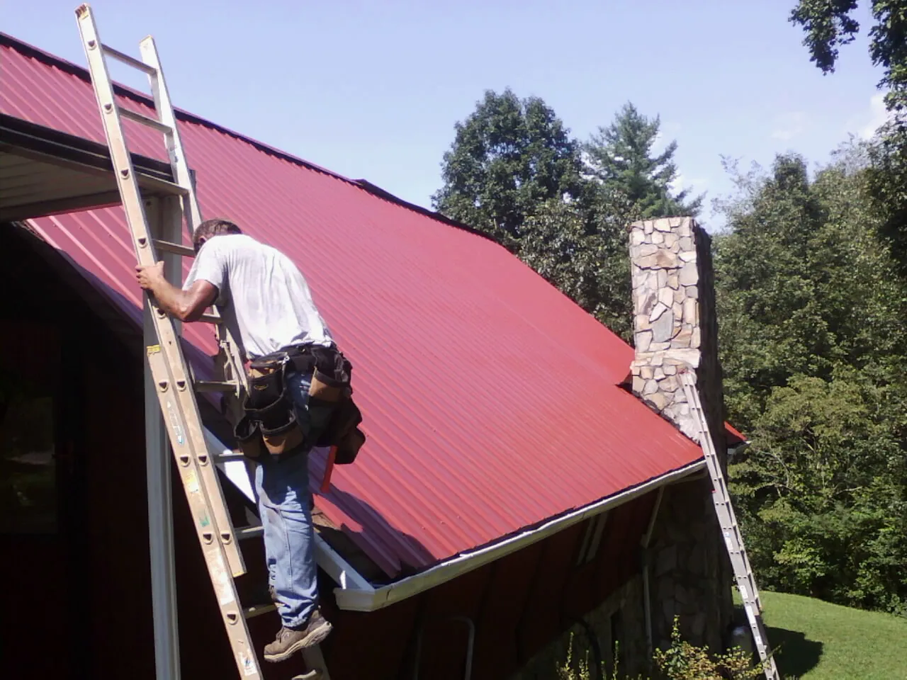 Roofer on a ladder beside a red metal roof and stone chimney — Soffit & Fascia Repair by Roofing Brothers in Clifton Springs